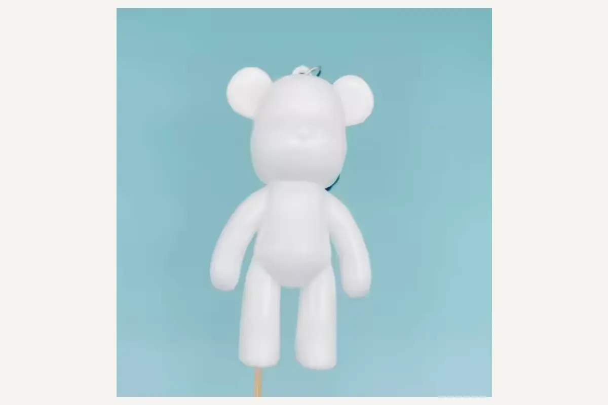 An unpainted block-shaped charm with projecting circular ears and long straight arms