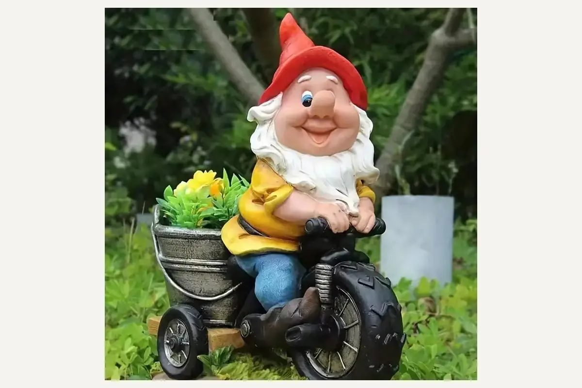 A smiling red hat garden gnome on a black wheeled vehicle with a flower bucket